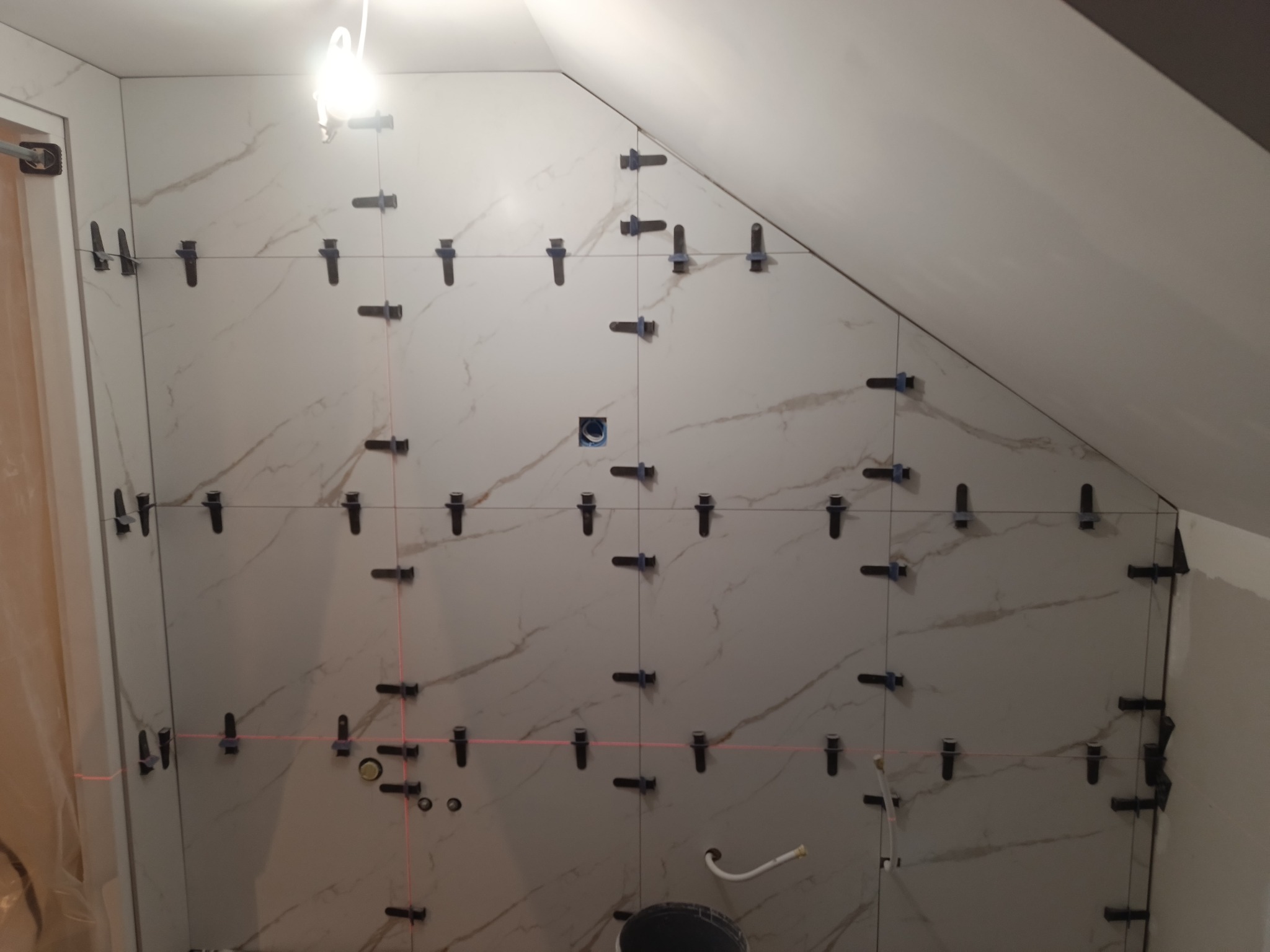 Ściana w trakcie układania płytek imitujących marmur, z użyciem systemu poziomowania płytek i lasera krzyżowego w łazience na poddaszu.