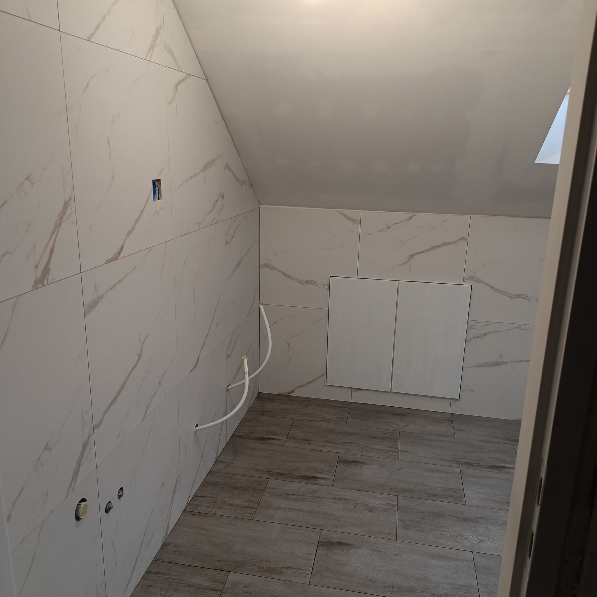 Wnętrze łazienki na poddaszu w trakcie prac wykończeniowych, widoczne białe płytki imitujące marmur na ścianach, drewnopodobna podłoga, instalacja wodna i otwór na gniazdko elektryczne.