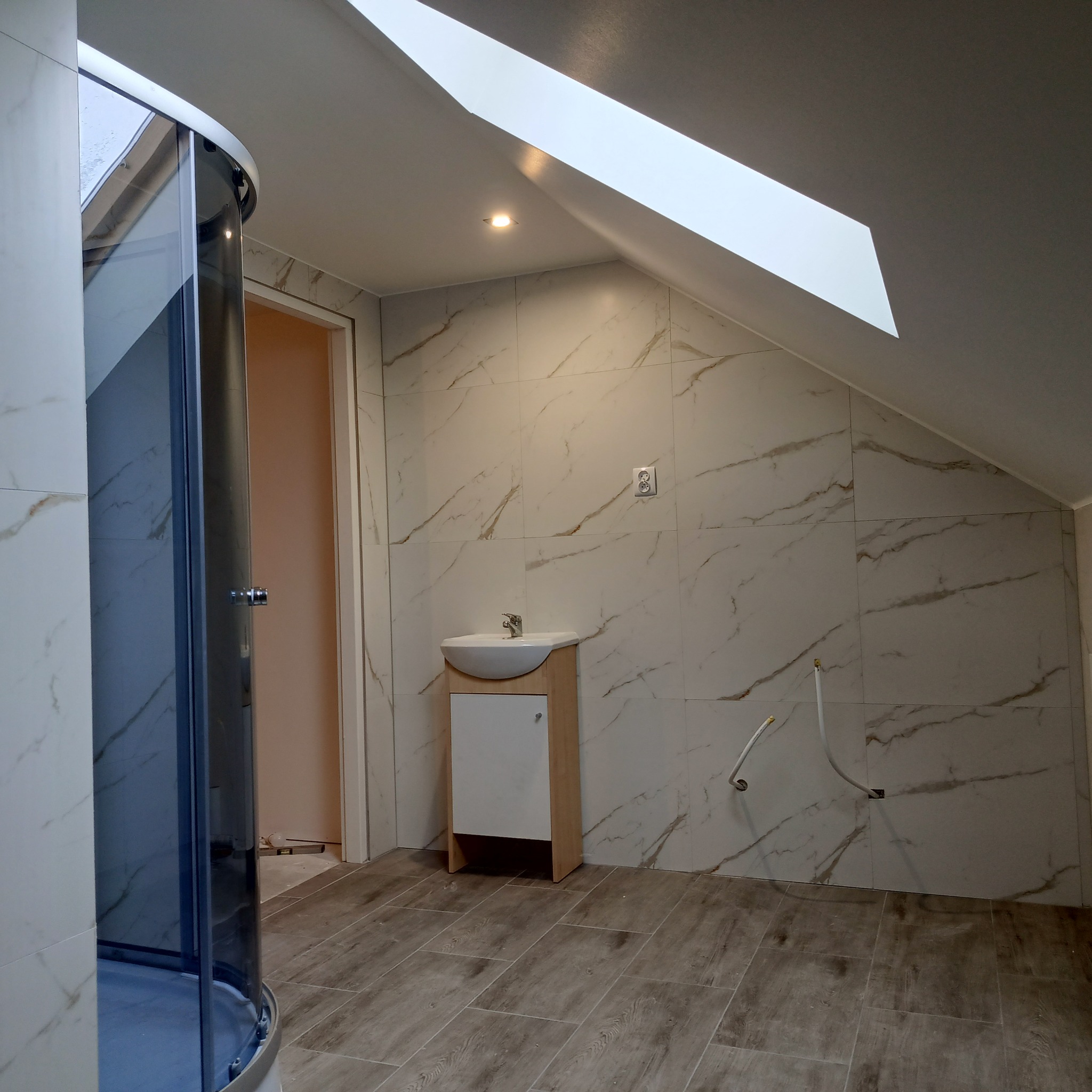 Wykończona łazienka na poddaszu w Tomaszowie Mazowieckim z kabiną prysznicową, umywalką z szafką i marmurowymi płytkami na ścianach. Widoczne wystające rury instalacji wodnej przed montażem armatury.