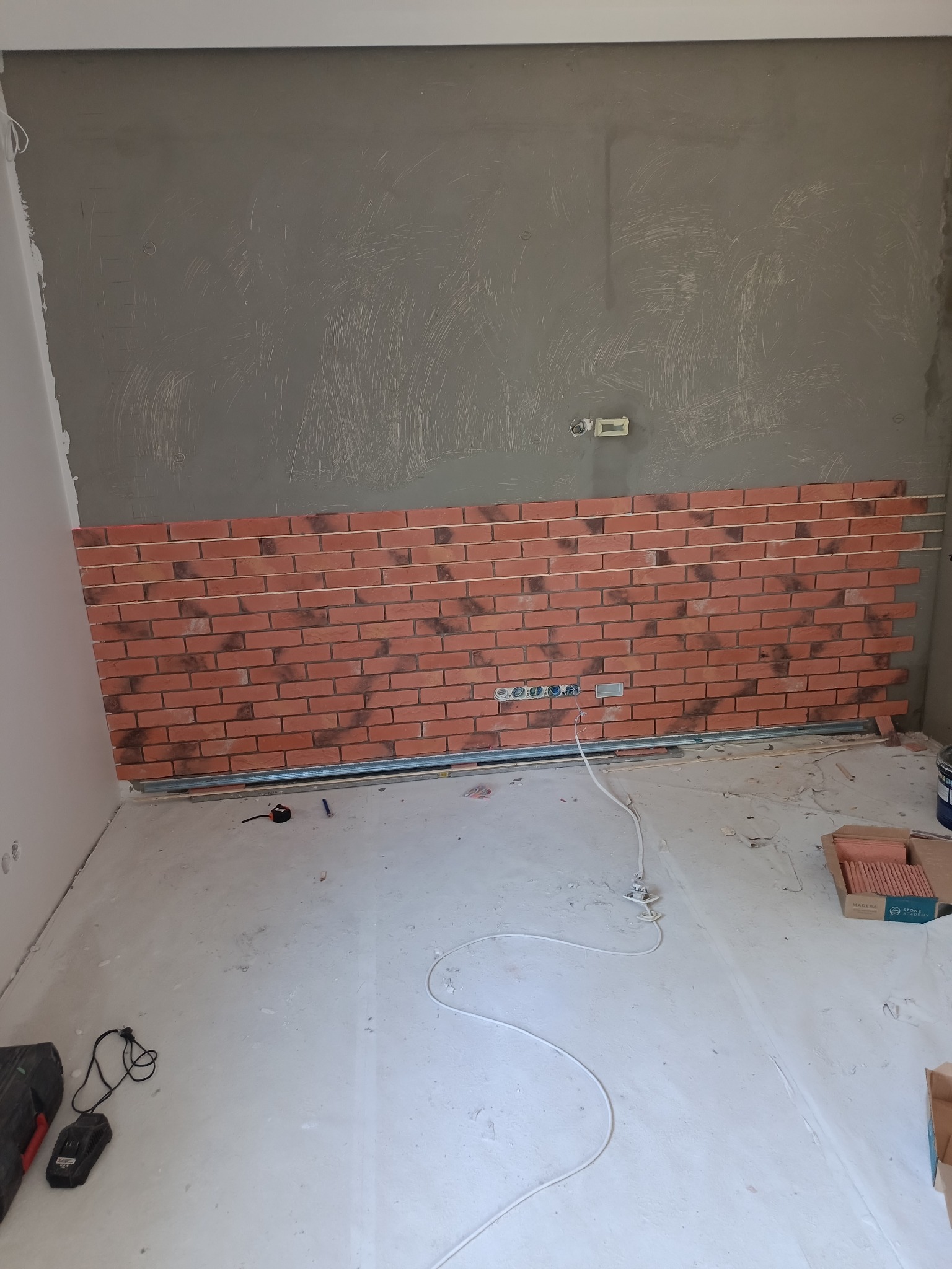 Montaż dekoracyjnej cegły na ścianie w trakcie prac wykończeniowych, widoczne profile montażowe, surowa ściana i podłoga, przewody elektryczne.