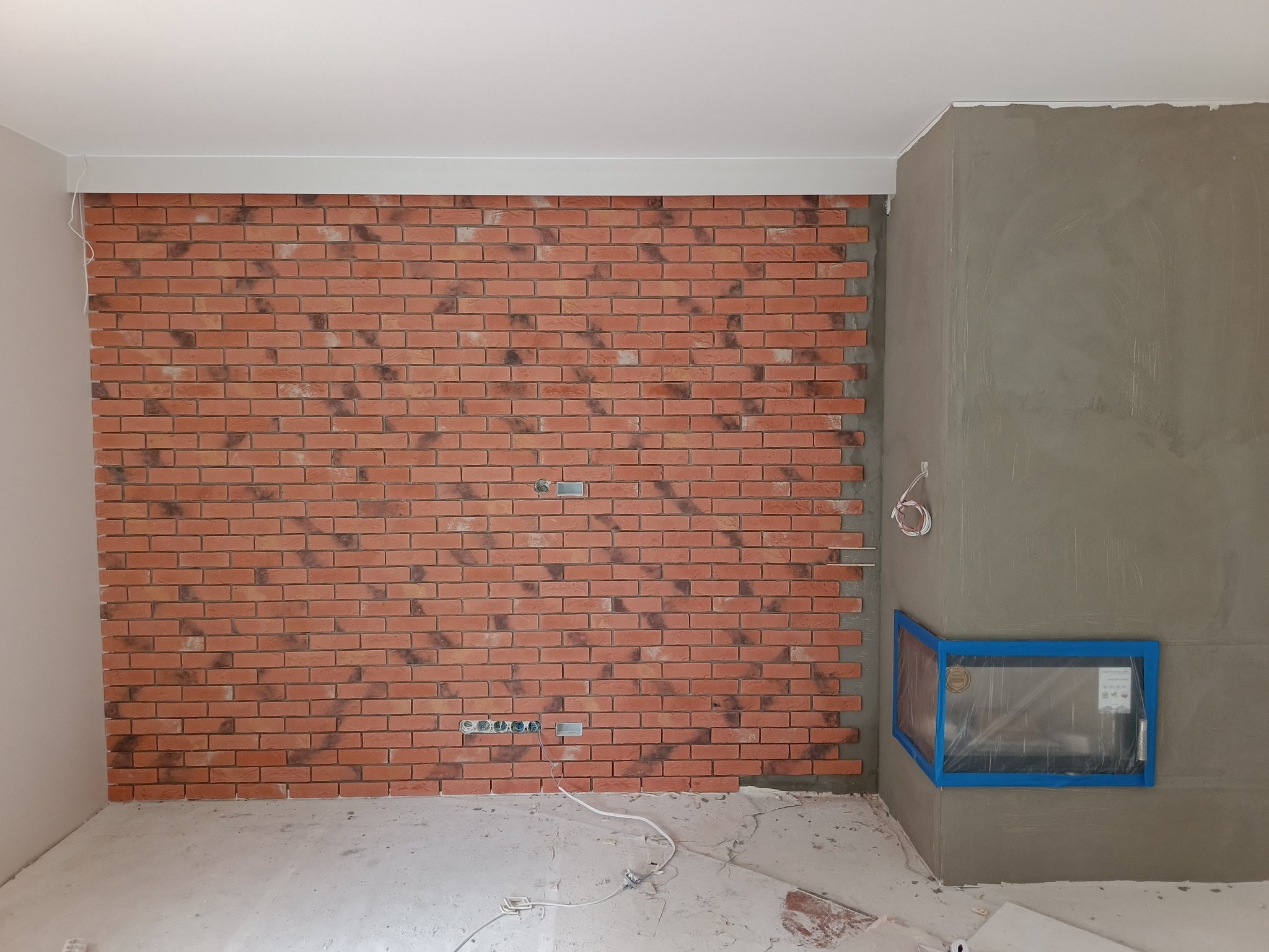 Ściana z dekoracyjnej cegły w trakcie montażu, widoczne metalowe szyny montażowe, szpachlowana ściana obok z zabezpieczonym niebieską taśmą kominkiem, surowa podłoga.