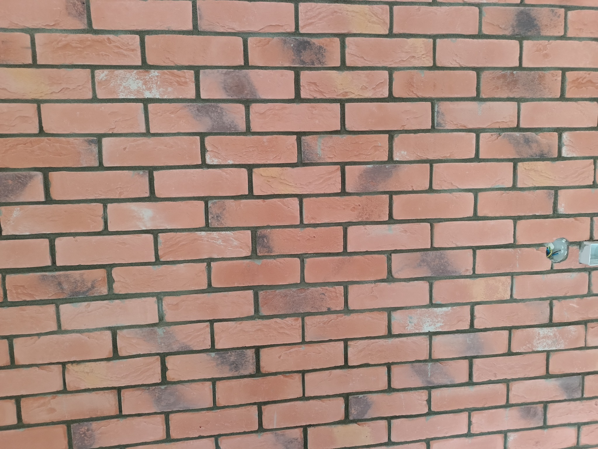 Ściana z dekoracyjnej cegły w odcieniach czerwieni i różu z ciemnoszarą fugą, widoczne gniazdko elektryczne z wystającymi przewodami.