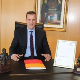 Mężczyzna w garniturze siedzi za biurkiem z folderem, certyfikatem w ramce i dwiema statuetkami. Na ścianie wisi obraz.