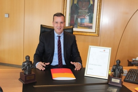 Mężczyzna w garniturze siedzi za biurkiem z folderem, certyfikatem w ramce i dwiema statuetkami. Na ścianie wisi obraz.