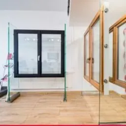 Wystawa okien PCV: czarne okno dwuskrzydłowe na szklanej podstawie obok okien drewnianych z klamkami, widoczne na jasnej podłodze.