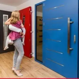 Dziewczynka niesiona na rękach przez kobietę na tle trzech różnych kolorystycznie drzwi wewnętrznych: niebieskich z poziomymi, srebrnymi wstawkami, czerwonych z szybą i czarnych. Jasna podłoga...