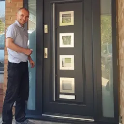 Szare drzwi wejściowe z czterema kwadratowymi przeszkleniami, zamontowane w domu z cegły, obok stoi uśmiechnięty mężczyzna w szarej koszulce i czarnych spodniach.