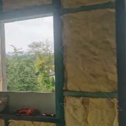 Ocieplenie ścian wełną mineralną w trakcie realizacji w Żywcu, widoczne drewniane ramy konstrukcyjne i sznurki mocujące izolację, z oknem w tle ukazującym zieleń.