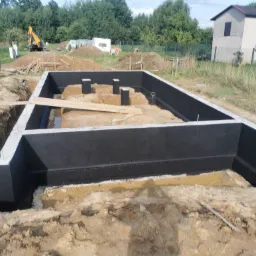 Wykop pod fundamenty z izolacją przeciwwilgociową, widoczny częściowo zalany wodą, z wykopaną ziemią i koparką w tle.