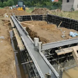 Wykop z zalewanymi betonem fundamentami, szalunki metalowe i drewniane, w tle koparka i budynek gospodarczy.