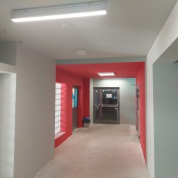 AGENCJA AGRAD - Korytarz po świeżym malowaniu z szarymi ścianami i intensywnie czerwonymi fragmentami, widoczne okno z luksferami i metalowe drzwi wejściowe, surowa podłoga przed wykończeniem.