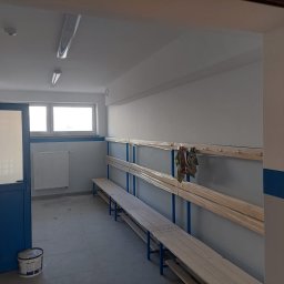 AGENCJA AGRAD - Wnętrze szatni po odświeżeniu: pomalowane ściany na szaro, niebieskie akcenty, drewniane ławki z wieszakami zabezpieczone folią, wiadro z farbą przy drzwiach.