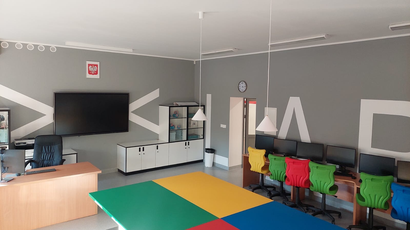 Wnętrze odnowionego biura z szarą ścianą ozdobioną białymi geometrycznymi wzorami i dużymi literami, kolorowymi krzesłami biurowymi i stołem z kolorowymi blatami, wyposażone w komputery i szafy.