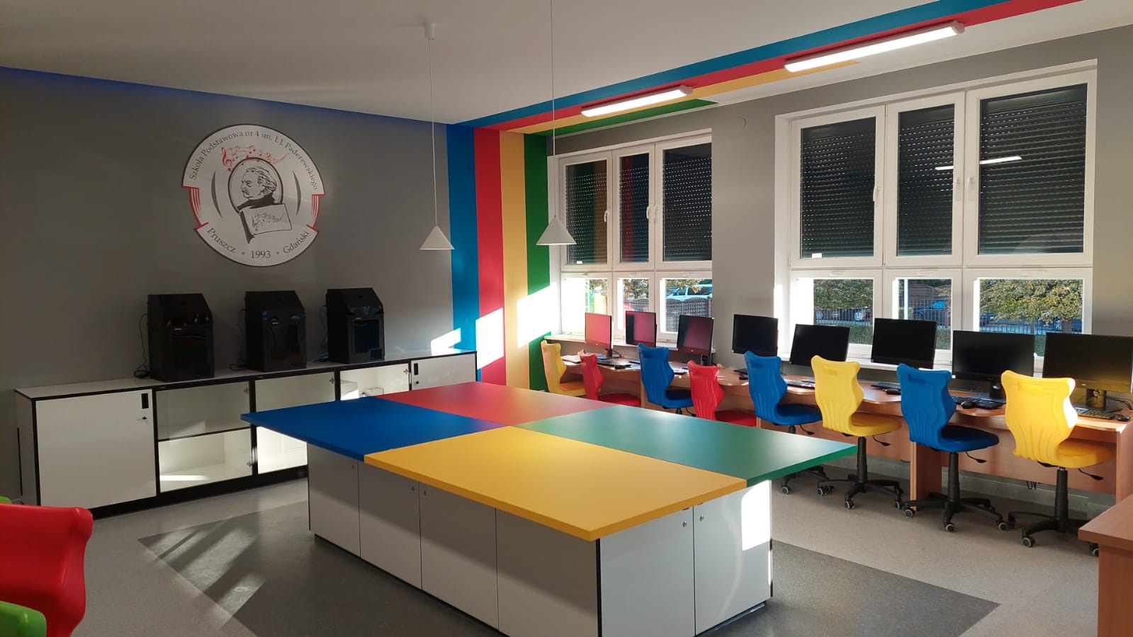 Kolorowa sala komputerowa w szkole podstawowej po remoncie, z biurkami i krzesłami w odcieniach niebieskiego, żółtego i czerwonego, oraz ścianami ozdobionymi pasami w kolorach tęczy i logo szkoły.