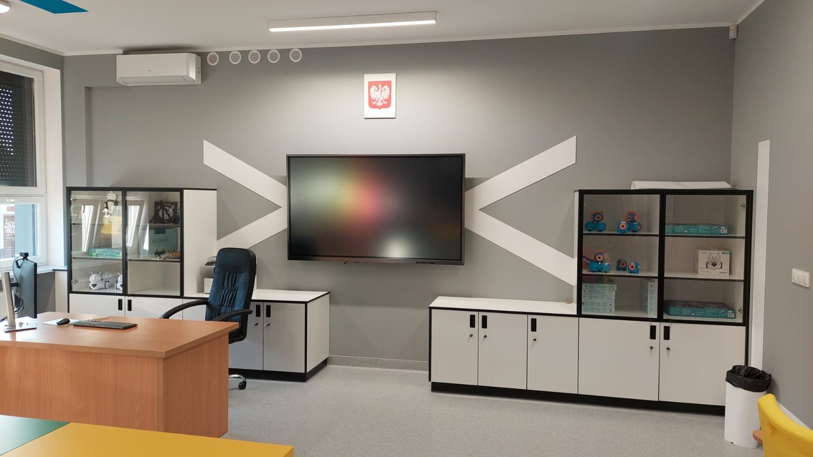 Jasne biuro z szarymi ścianami, biurkiem z drewna, szafkami i dużym ekranem interaktywnym na ścianie z dekoracyjnymi białymi pasami w kształcie litery X.