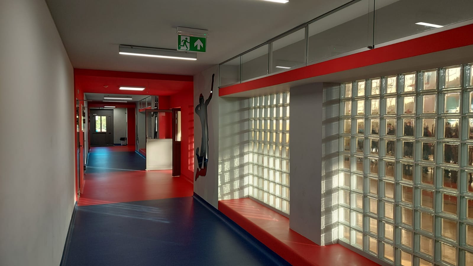 Korytarz w budynku biurowym z czerwonymi akcentami, ścianą z luksferów i grafiką sportowca na ścianie.