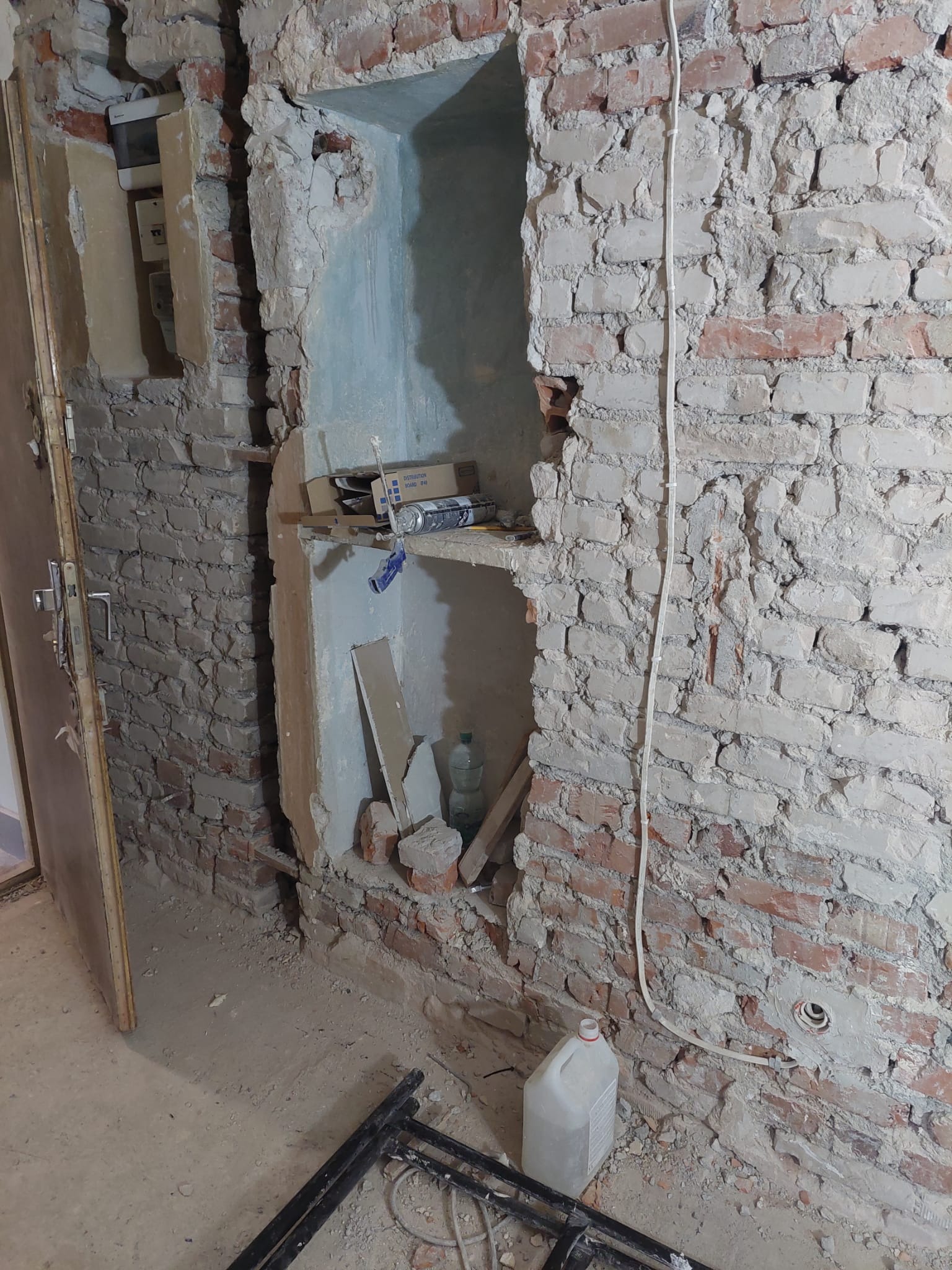 Surowa ściana z czerwonej cegły z wyłamanymi wnękami, widoczne przewody elektryczne, resztki gruzu i narzędzia na podłodze, drzwi w tle. Etap przygotowawczy do prac wykończeniowych.