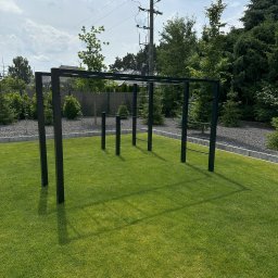 KPE & MP DESIGN SP. Z O.O. - Metalowa konstrukcja do ćwiczeń na trawniku, z elementami drabinki i drążków, otoczona zielenią. Widok na ogród i słupy energetyczne w tle.