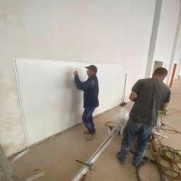 KPE & MP DESIGN SP. Z O.O. - Dwóch monterów instaluje metalowe panele ścienne w jasnym pomieszczeniu. Jeden z pracowników przytwierdza panel, drugi stoi obok z narzędziami.