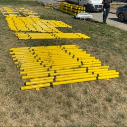 KPE & MP DESIGN SP. Z O.O. - Żółte, metalowe słupki ogrodzeniowe z czarnymi pasami, ułożone na trawie w rzędach. W tle biały bus i osoba trzymająca telefon.