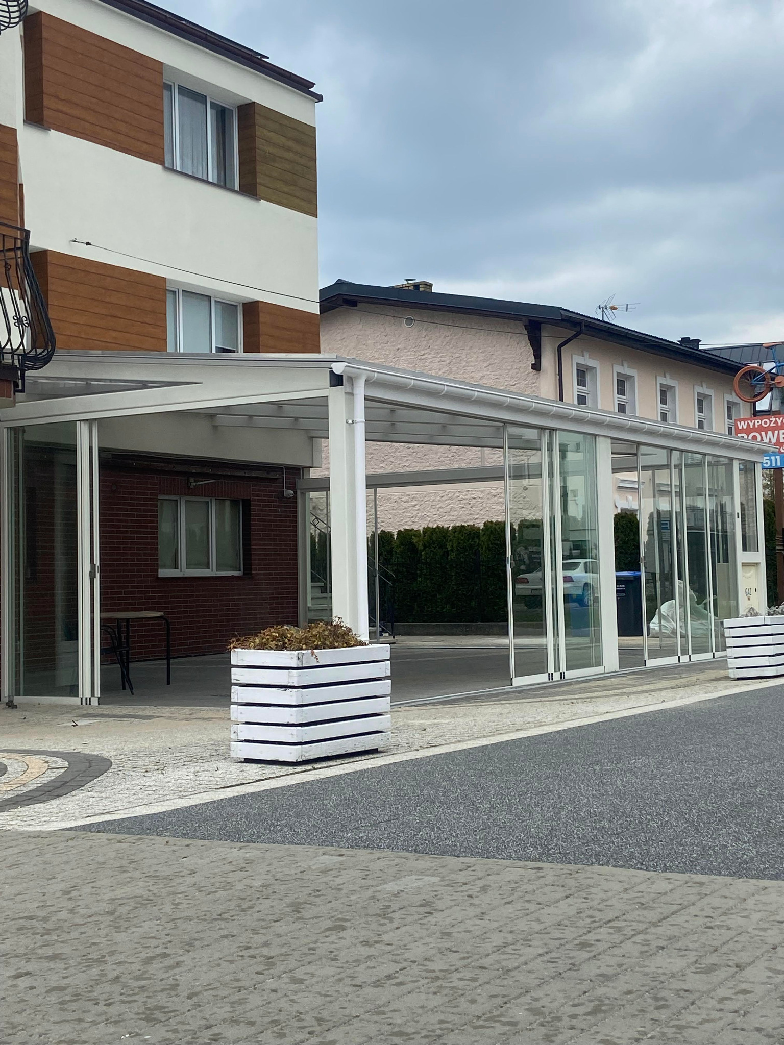 Zadaszenie tarasu z przesuwnymi szklanymi panelami, wsparte na białych słupach, z widocznym stołem i krzesłami wewnątrz. Białe donice z roślinami na zewnątrz.