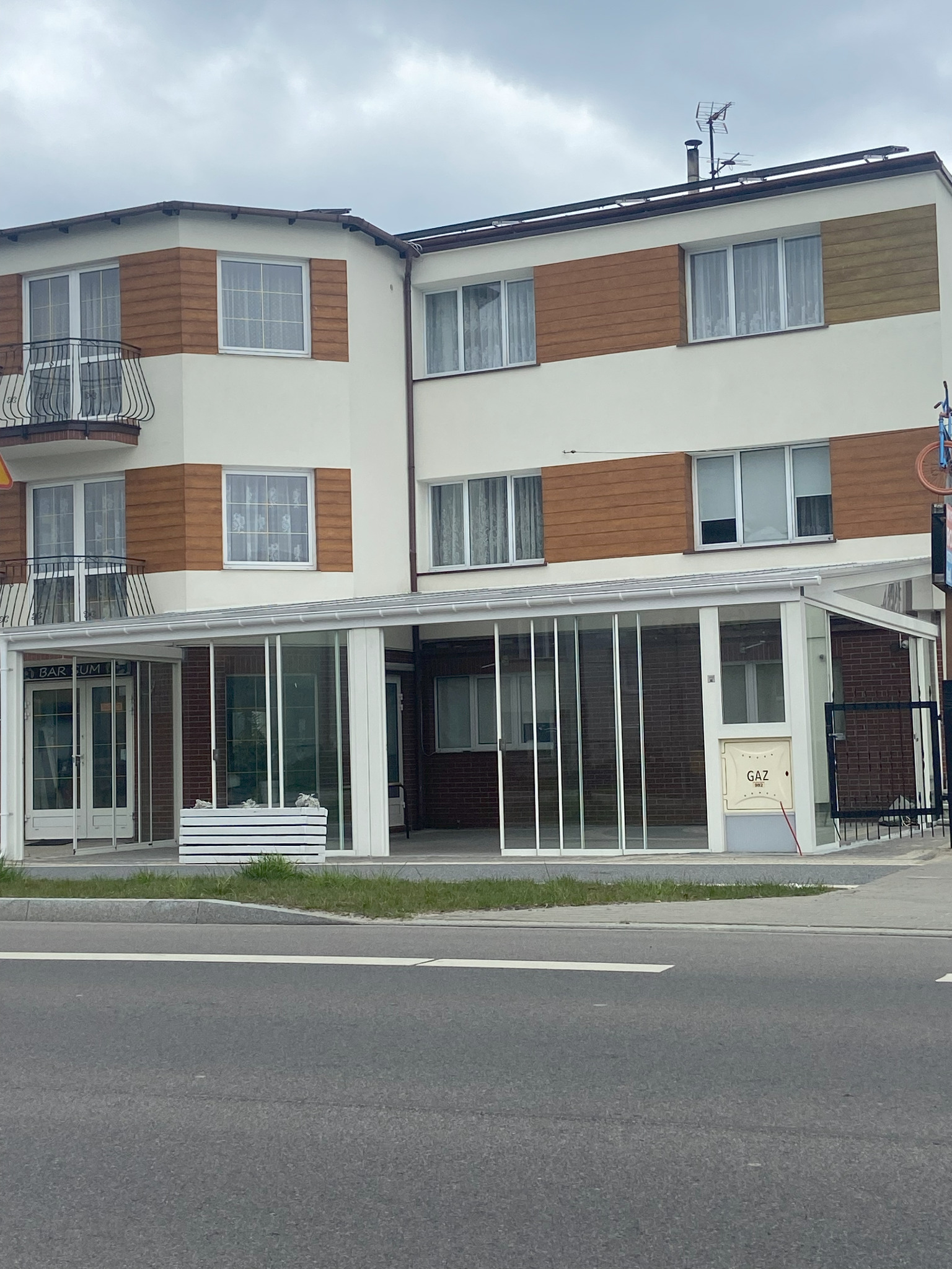 Budynek z balkonami i przeszklonym wejściem, widok z ulicy. Elementy balustrad balkonowych wykonane z metalu. Widoczna skrzynka na kwiaty.