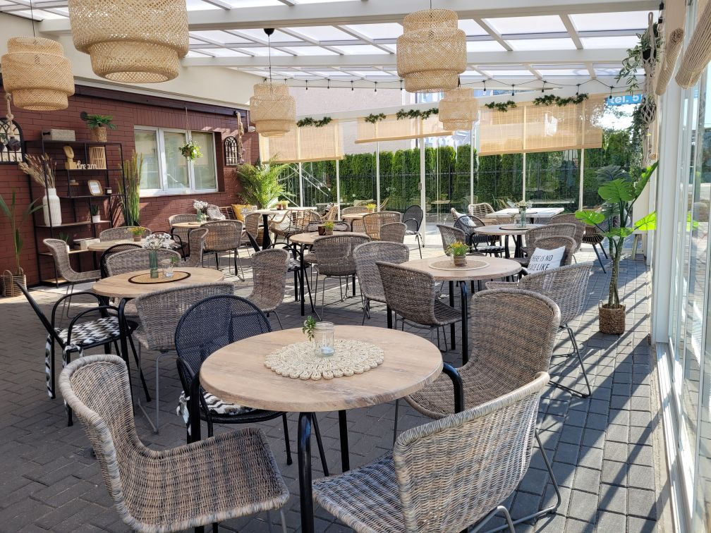 Zadaszony taras z rattanowymi meblami i lampami. Widoczne stoły z krzesłami, dekoracje roślinne i oświetlenie girlandowe. Przeszklona konstrukcja zapewnia dostęp światła.