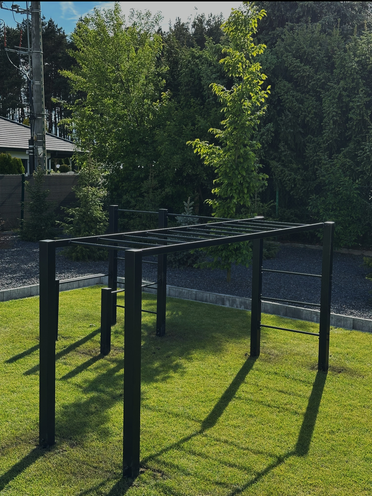 Czarna, metalowa konstrukcja do ćwiczeń na trawniku, w tle ogród z drzewami i krzewami. Minimalistyczny design, nowoczesny wygląd, równomierne oświetlenie słoneczne.