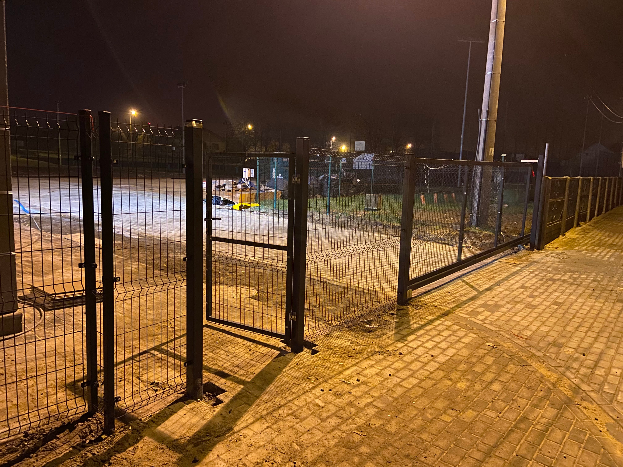 Otwarta, przesuwna brama panelowa z widocznym ogrodzeniem na terenie inwestycji oświetlonym lampami ulicznymi. Nocna sceneria, kostka brukowa.
