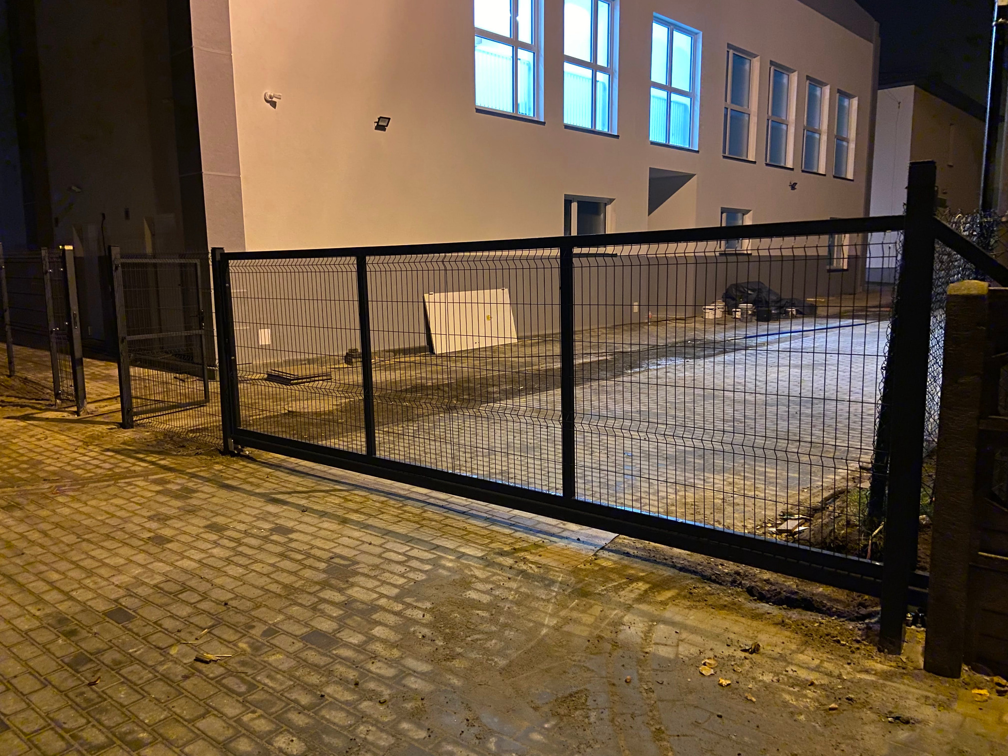 Nowoczesna, przesuwna brama panelowa z czarną ramą, widoczna na tle budynku z oświetlonymi oknami. Brama zamontowana na kostce brukowej, oświetlona światłem ulicznym.