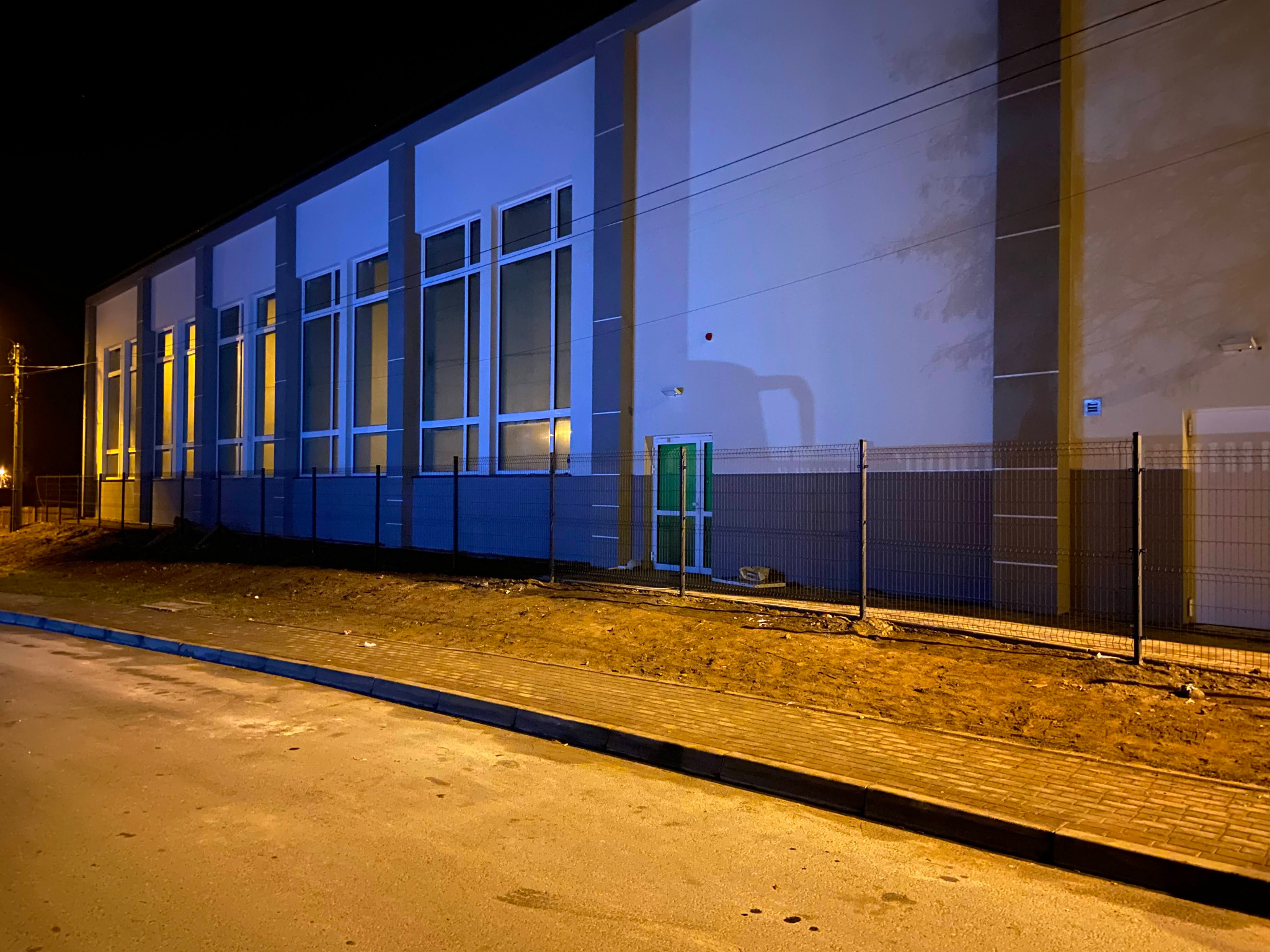 Nowoczesny budynek przemysłowy z ogrodzeniem panelowym w kolorze grafitowym, oświetlony nocą. Widoczna zielona brama wejściowa i fragment chodnika.