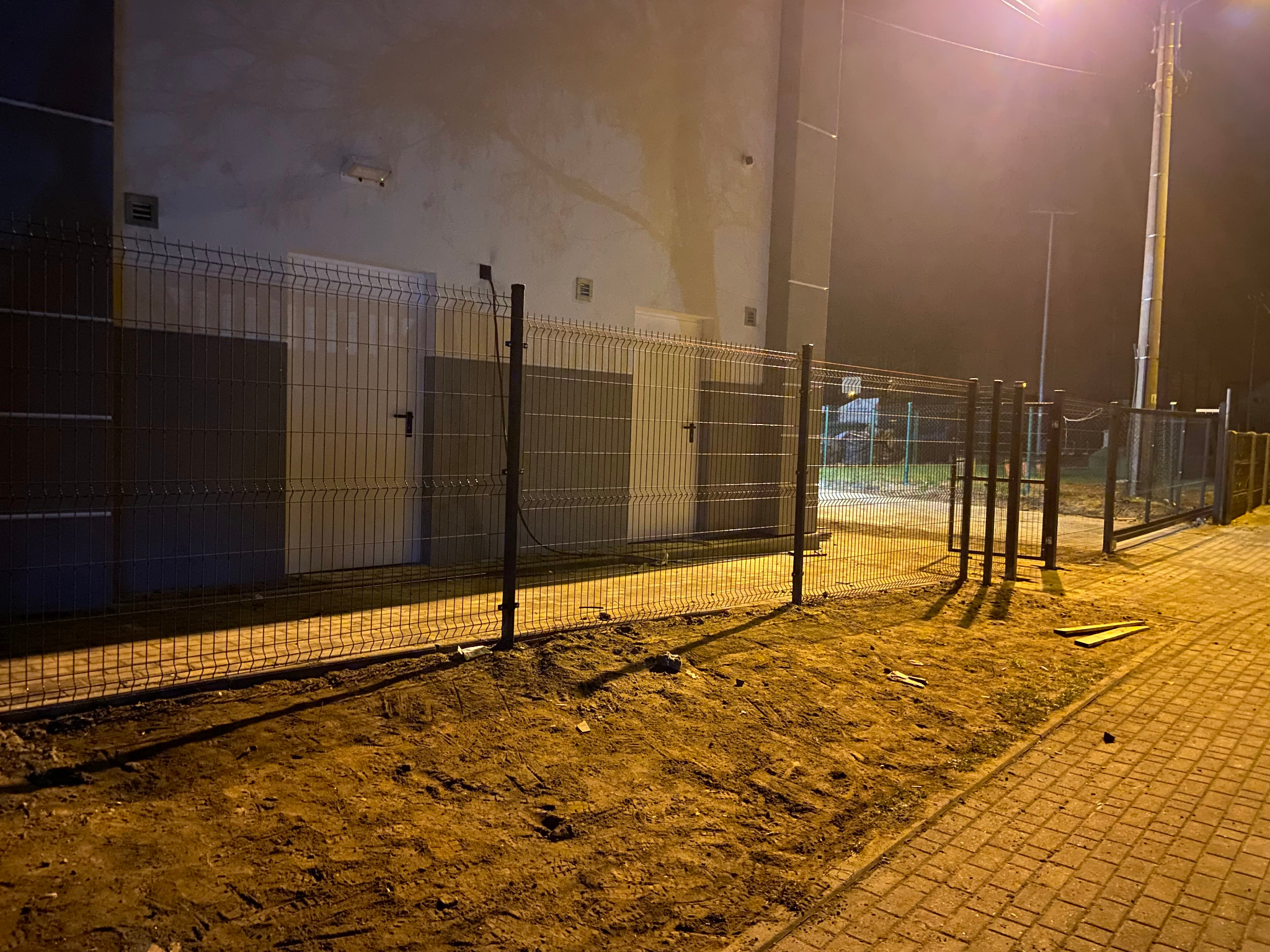 Częściowo zamontowane ogrodzenie panelowe z metalową furtką, oświetlone nocą. Widoczne słupki i panele. Tło stanowi budynek i teren z ziemią. Elementy montażowe na ziemi.