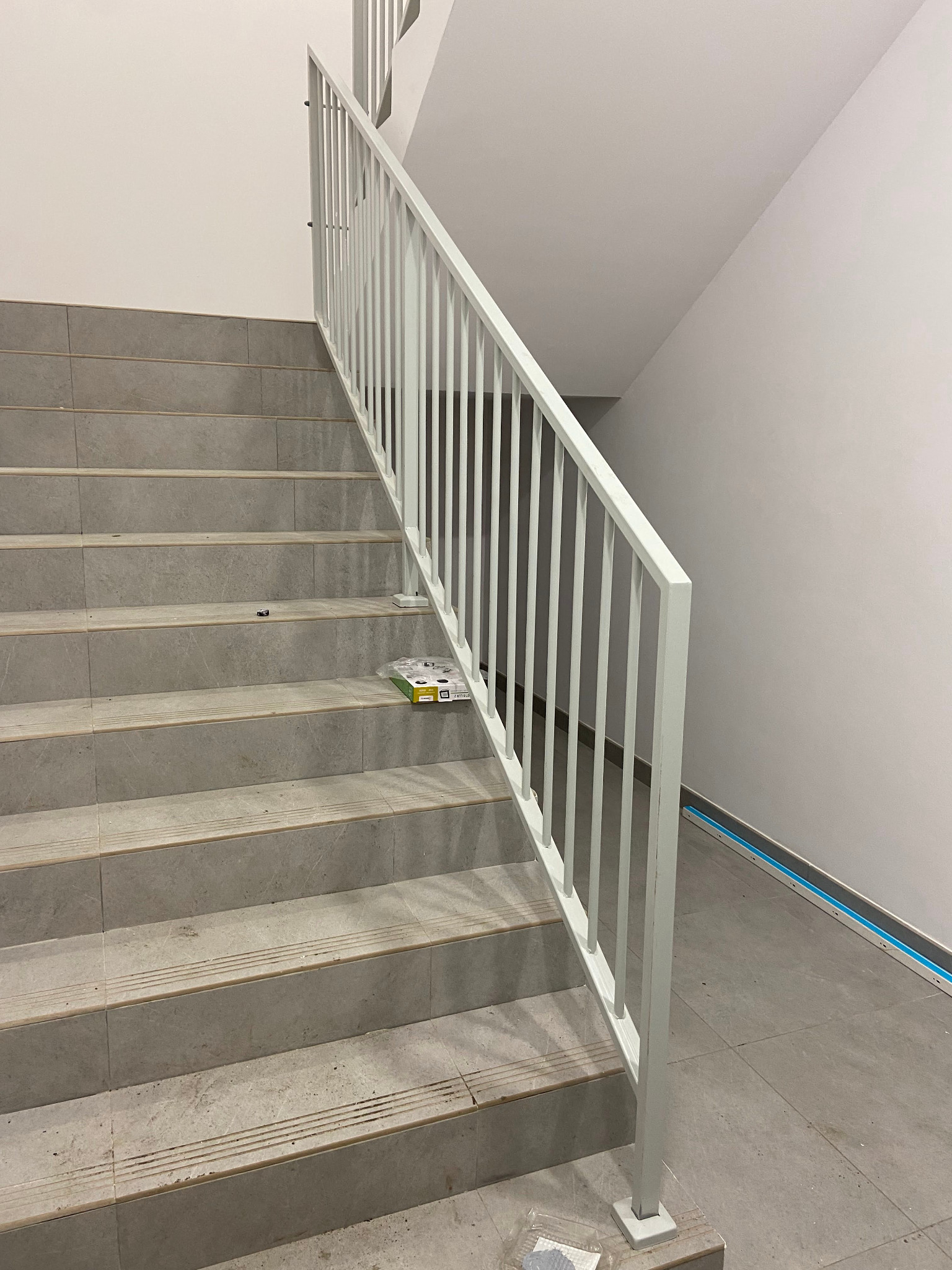 Biała balustrada metalowa na schodach z szarych płytek. Widoczne elementy montażowe na stopniach. Minimalistyczny design, jasne wnętrze.