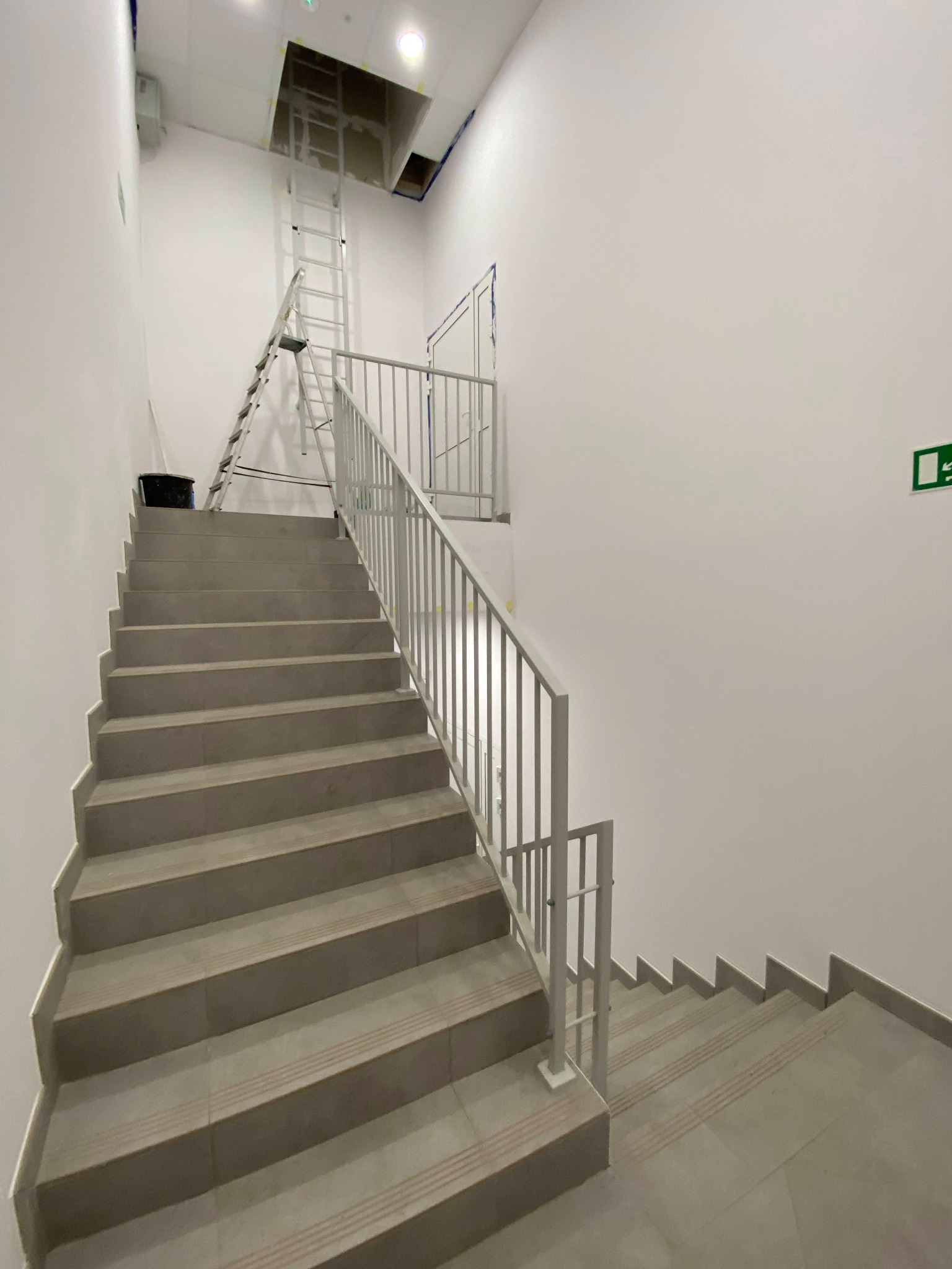 Jasne schody wyłożone płytkami z metalową balustradą, drabina oparta o sufit. Widoczny znak ewakuacyjny. Wykończenie w minimalistycznym, nowoczesnym stylu.