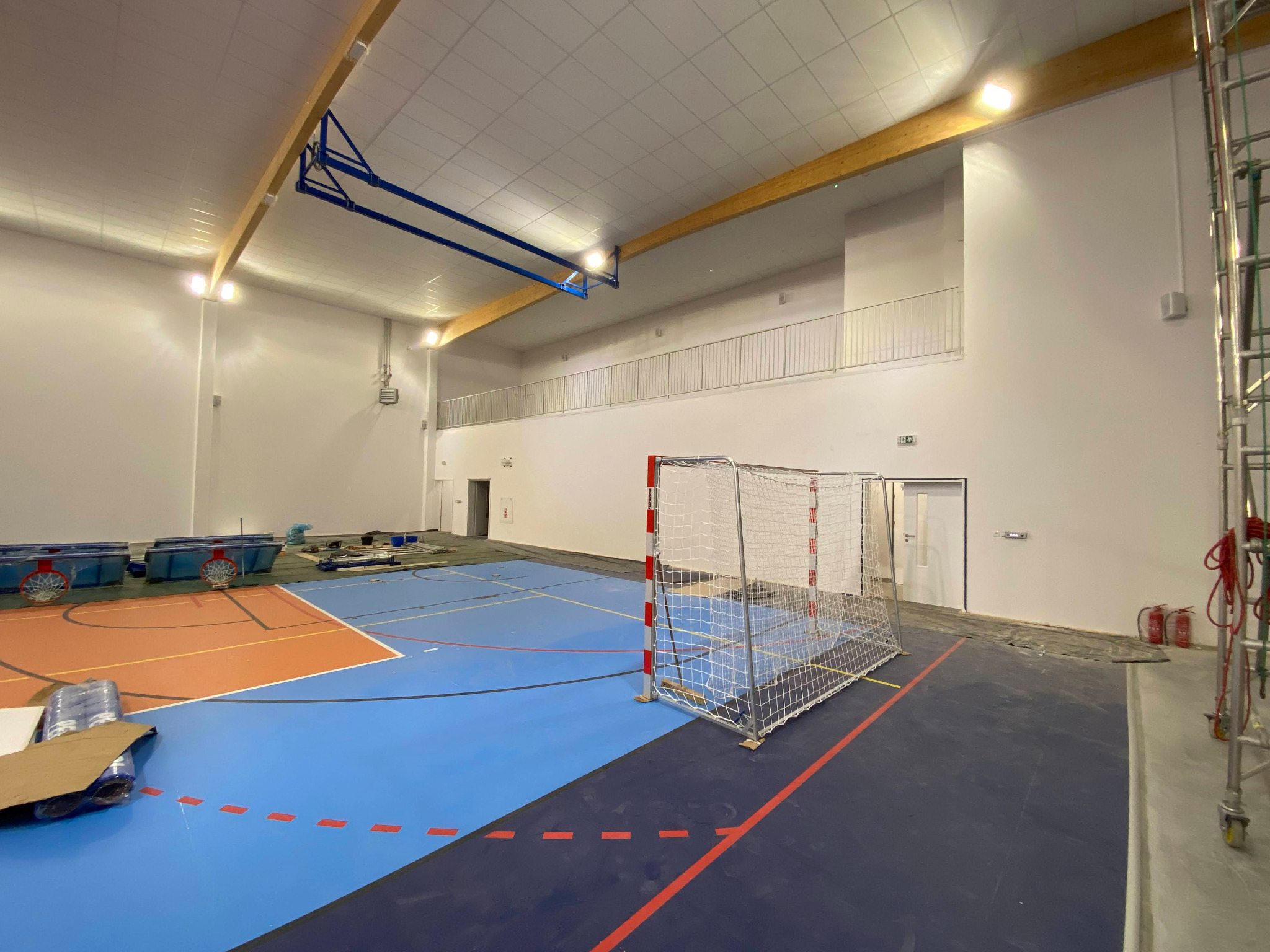 Hala sportowa w trakcie budowy: widoczny parkiet w kolorach niebieskim i pomarańczowym, bramka do piłki ręcznej, rusztowania i elementy konstrukcyjne pod sufitem.