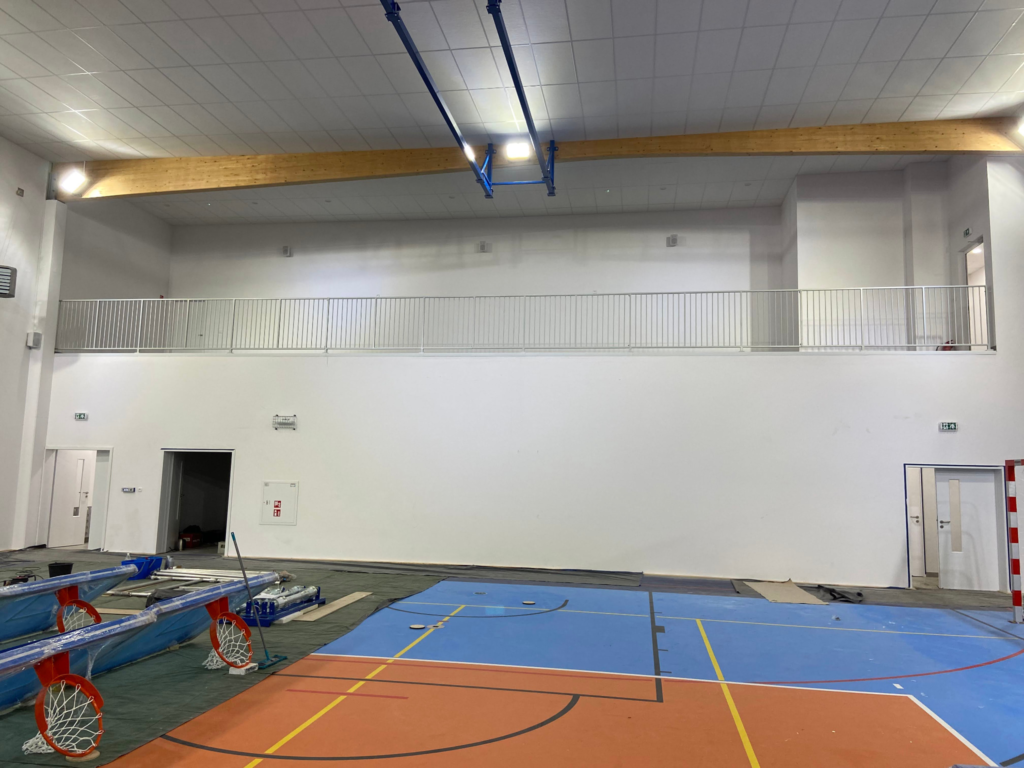 Wnętrze hali sportowej z metalową balustradą na antresoli. Na podłodze leżą elementy koszykówki, co sugeruje trwający montaż lub przygotowanie do użytku.
