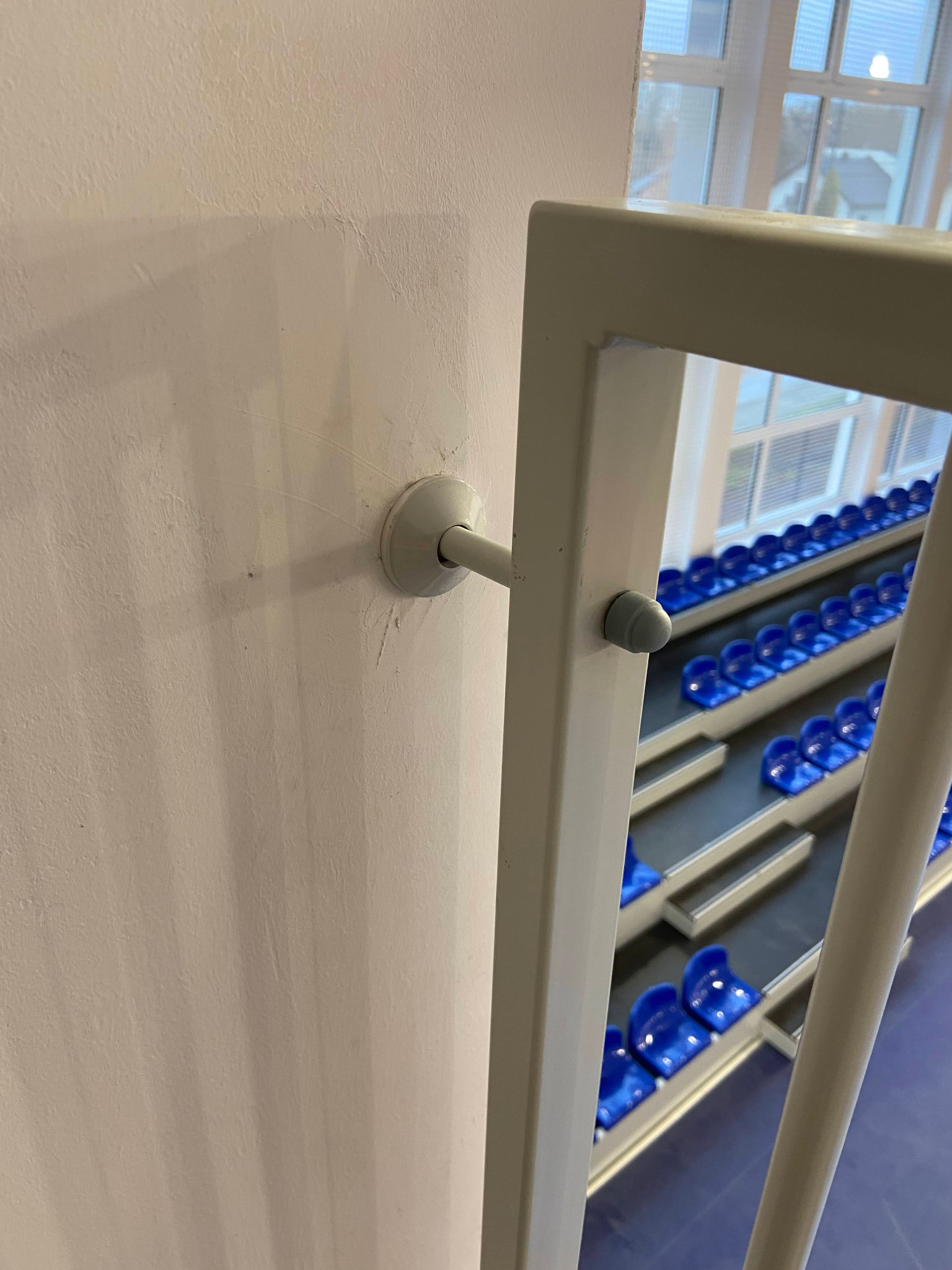 Szczegół mocowania metalowej balustrady schodowej na ścianie, z widokiem na rzędy niebieskich krzesełek na trybunach w tle. Jasne tło, neutralna kolorystyka.