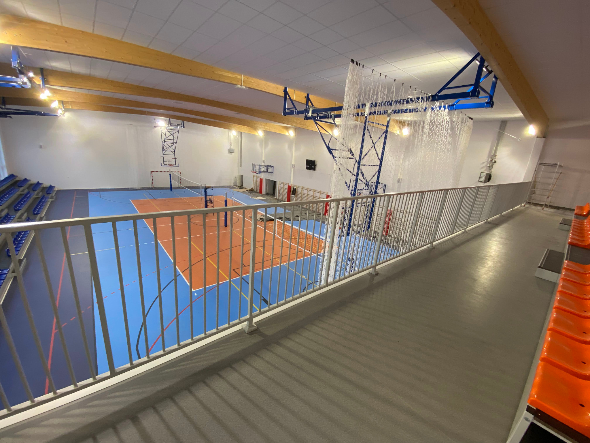 Widok z góry na halę sportową z balustradą. Parkiet z liniami boisk do różnych dyscyplin, bramki i siatki. Widoczne trybuny i drewniane belki stropowe.