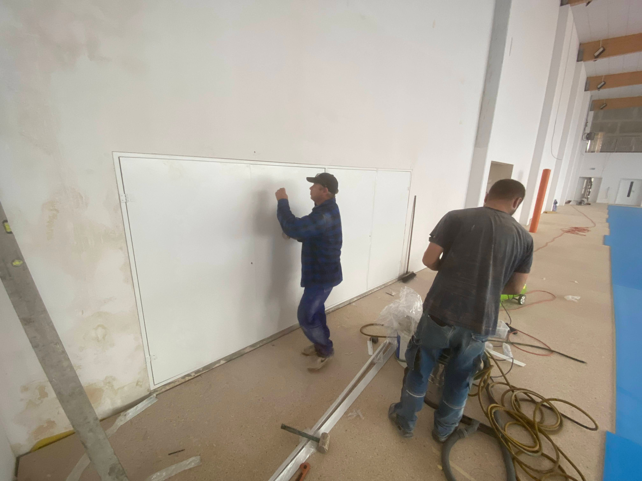 Dwóch monterów instaluje metalowe panele ścienne w jasnym pomieszczeniu. Jeden z pracowników przytwierdza panel, drugi stoi obok z narzędziami.