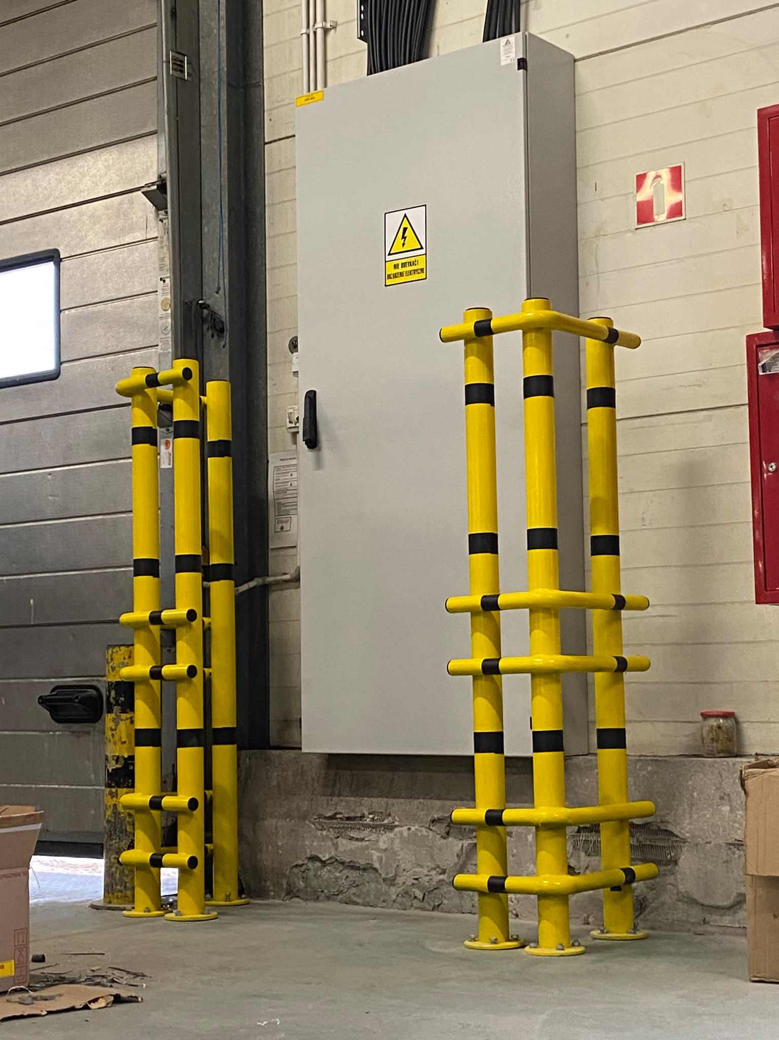 Żółte bariery ochronne z czarnymi pasami, zabezpieczające przed uszkodzeniami w hali przemysłowej, widoczne przy szarej szafie elektrycznej i metalowej bramie.