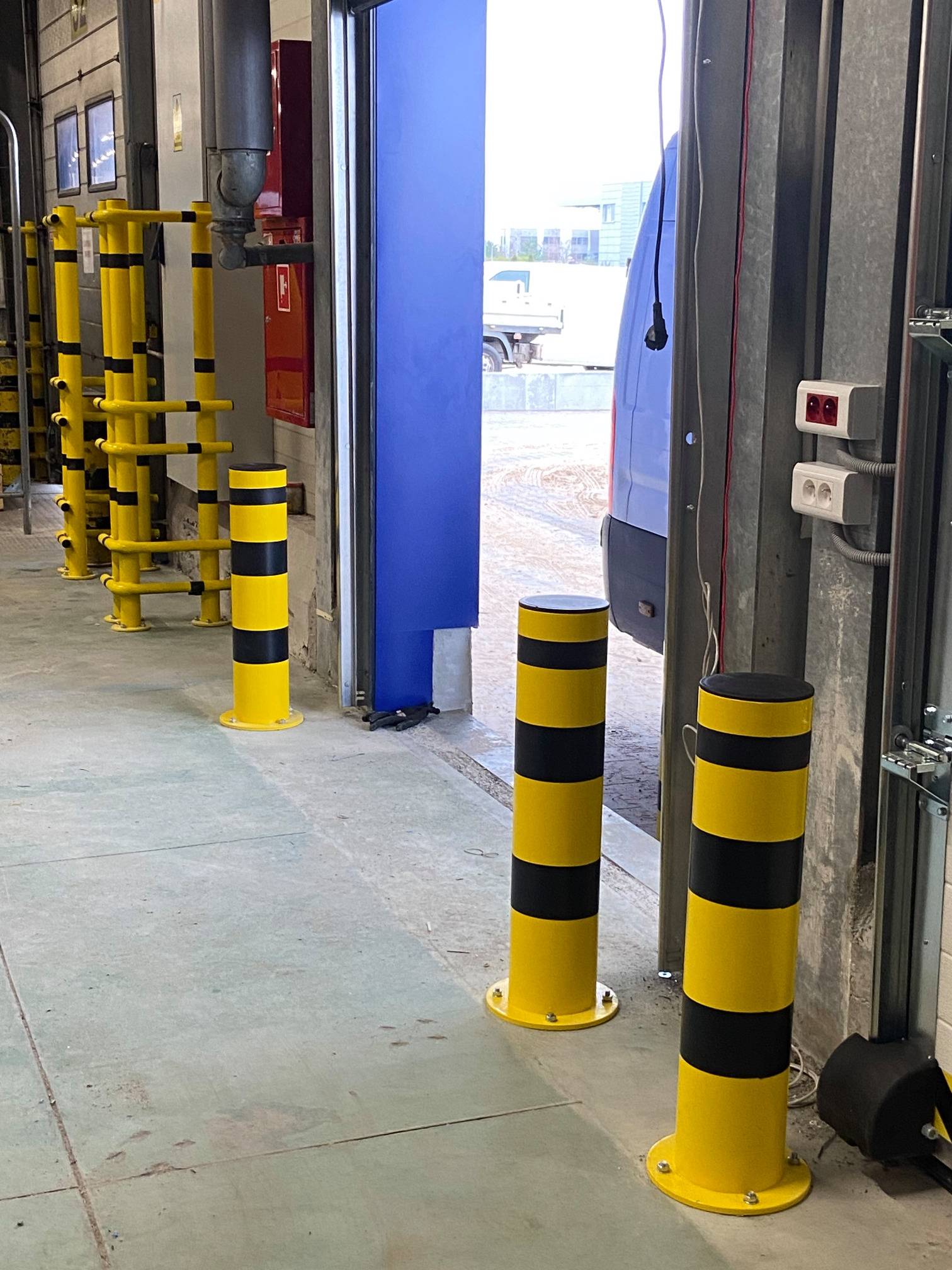 Żółto-czarne słupki ochronne w hali przemysłowej, zamontowane przy bramie wjazdowej. Widoczne ślady użytkowania na podłodze i elementach konstrukcyjnych. Otwarta brama w tle.