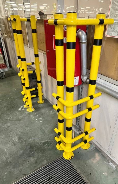 Żółte, metalowe słupki ochronne z czarnymi pasami, zamontowane na podłodze w hali. Słupki zabezpieczają czerwoną skrzynkę hydrantową i rurę. Widoczna kratka ściekowa.