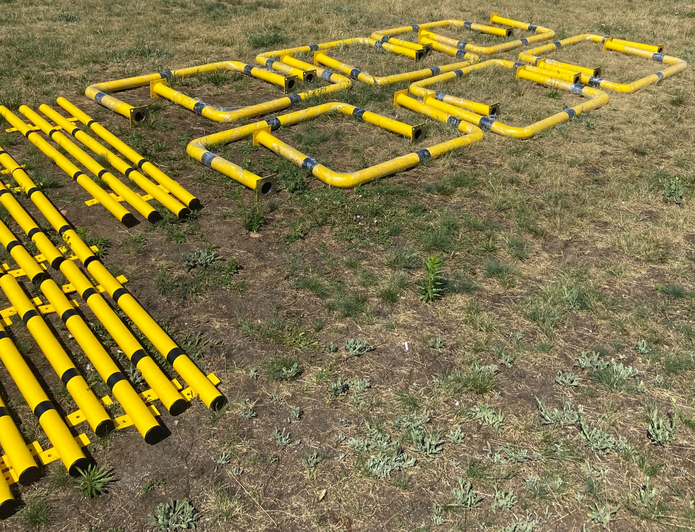 Żółte, metalowe elementy konstrukcyjne z czarnymi akcentami, leżące na trawie i ziemi. Różne kształty: rury i kwadratowe ramy. Przygotowane do montażu ogrodzenia lub balustrady.