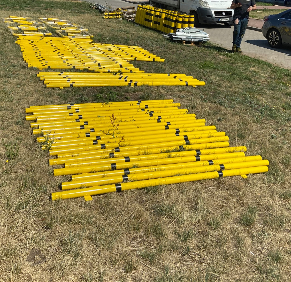 Żółte, metalowe słupki ogrodzeniowe z czarnymi pasami, ułożone na trawie w rzędach. W tle biały bus i osoba trzymająca telefon.