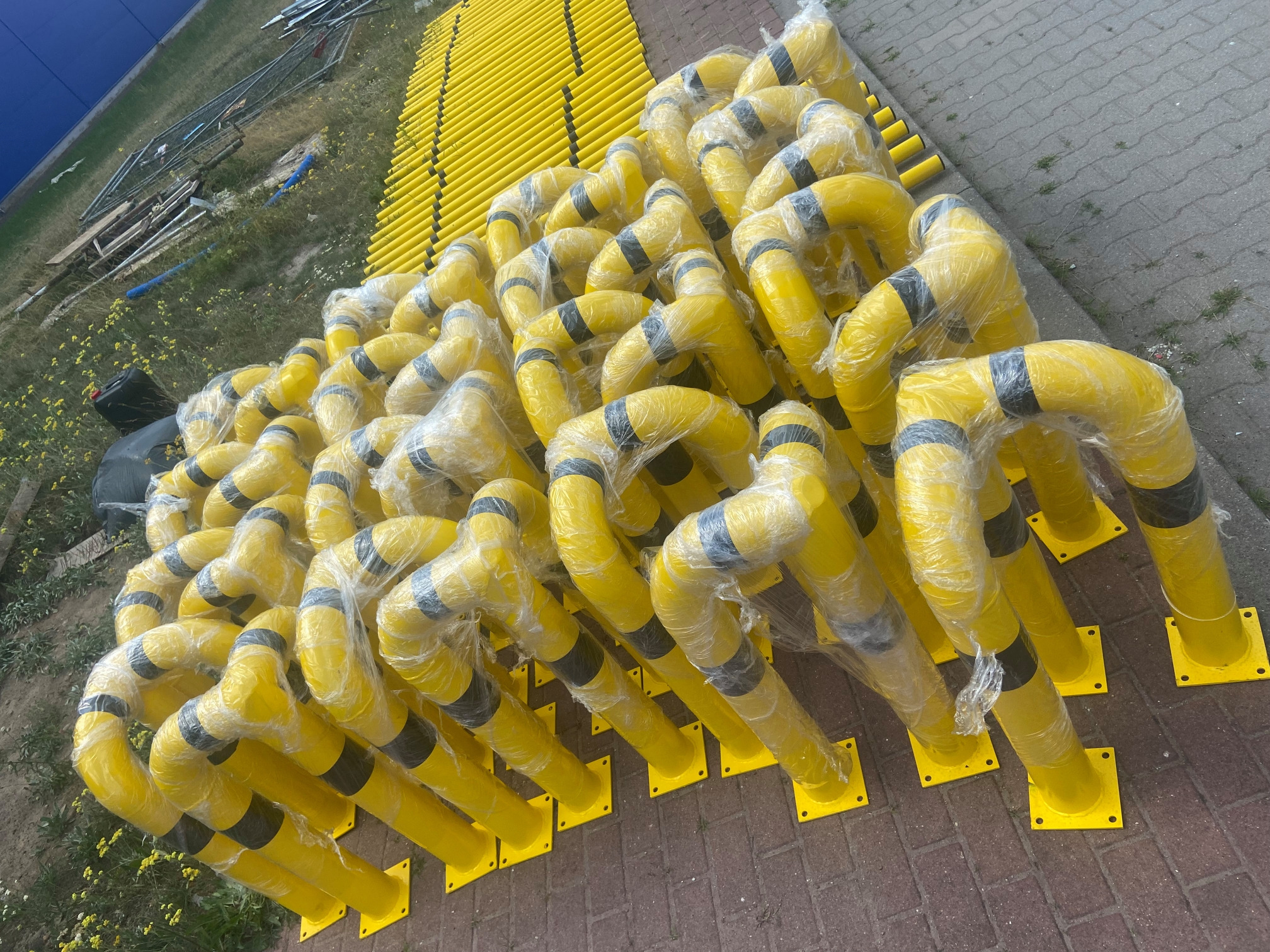 Żółte, metalowe słupki zabezpieczające z czarnymi pasami, owinięte folią, ułożone na kostce brukowej. W tle widoczne inne elementy konstrukcyjne i roślinność.