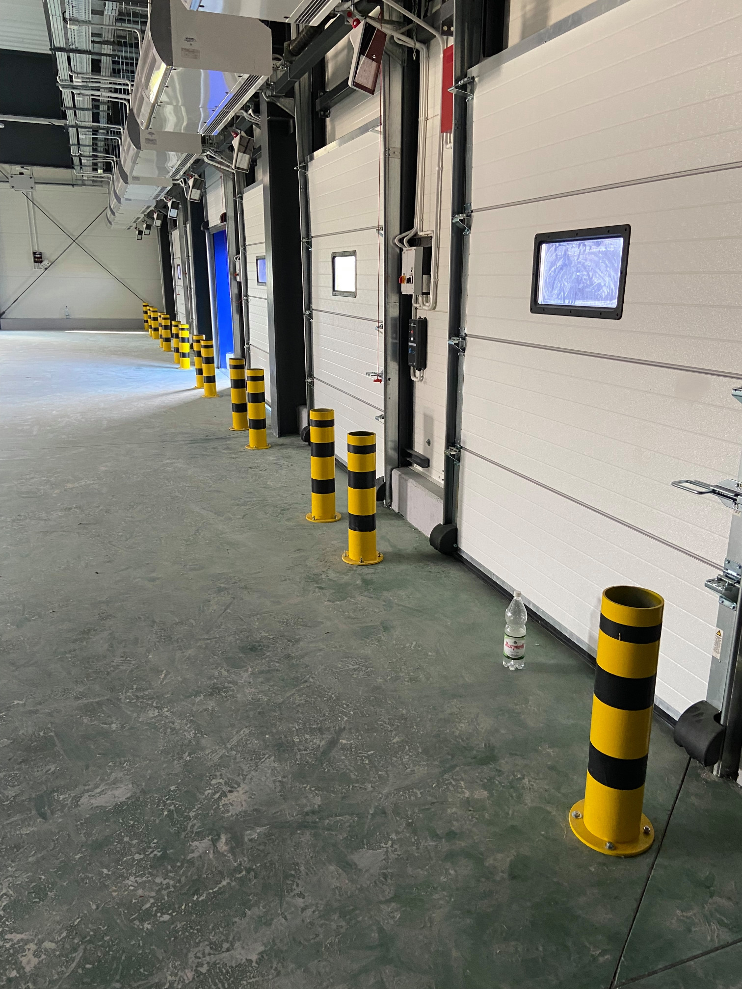 Rząd białych bram segmentowych z oknami w hali magazynowej w Białych Błotach, oddzielonych żółto-czarnymi słupkami ochronnymi.