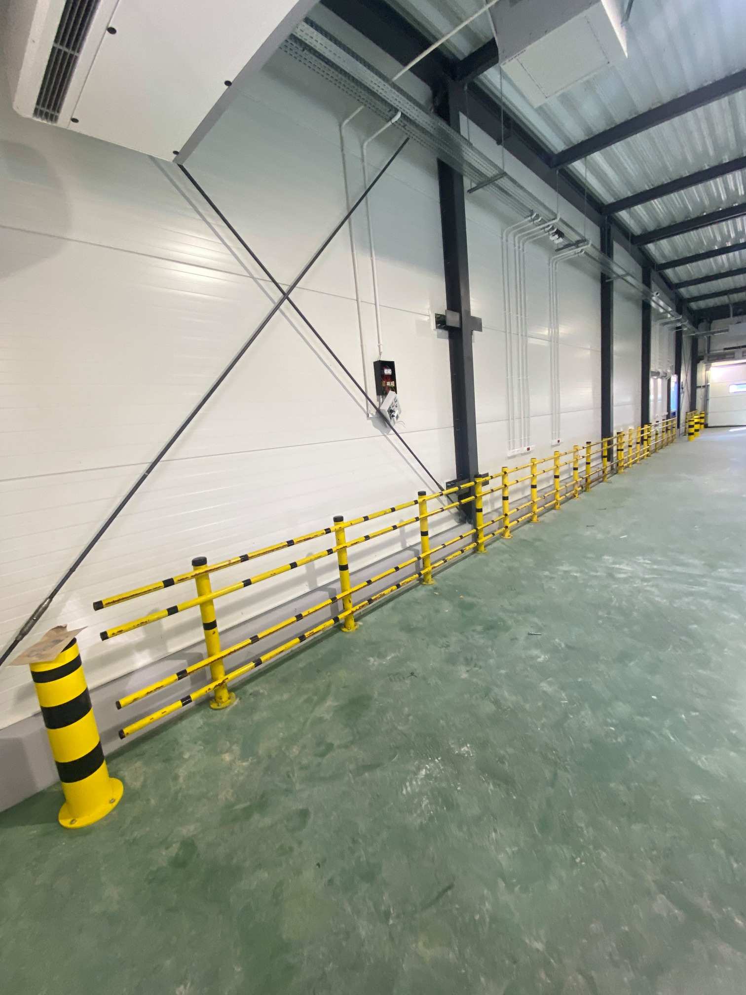 Żółto-czarne barierki ochronne wzdłuż ściany hali przemysłowej. Metalowa konstrukcja dachu i białe ściany. Ochrona maszyn i pieszych w obiekcie.