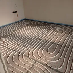 Białe rury ogrzewania podłogowego rozłożone na czerwonej siatce, widoczne mocowania, fragment ściany z otworem instalacyjnym w rogu pomieszczenia.