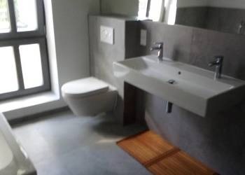 Nowoczesna łazienka z podwieszaną toaletą i umywalką, szare płytki, drewniana mata podłogowa, widok z boku.