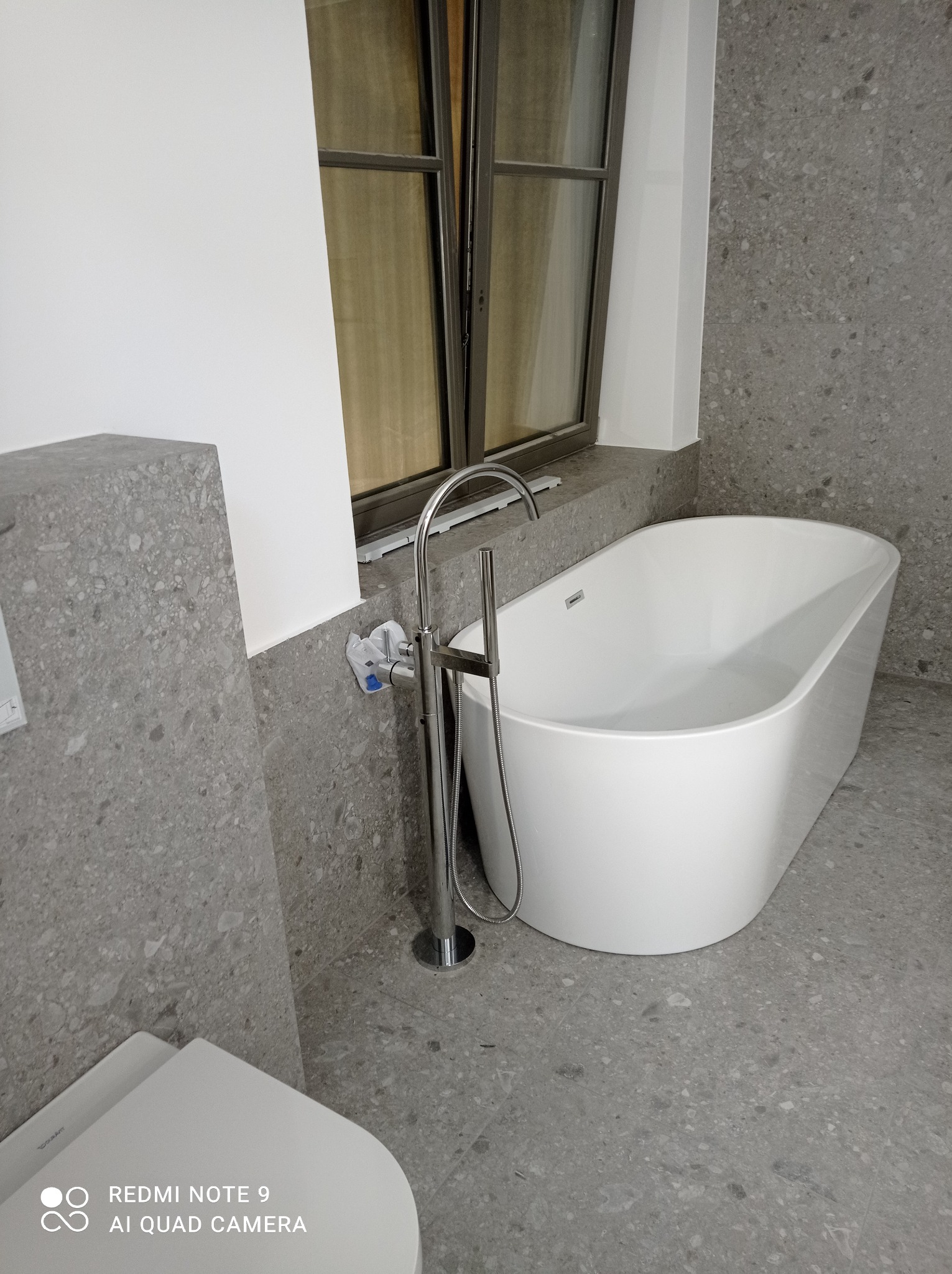 Nowoczesna, wolnostojąca wanna z chromowaną baterią podokienną, na tle szarej, lastrykowej posadzki i okładziny ściennej. Częściowo widoczna toaleta. Zabezpieczenie na zaworze.